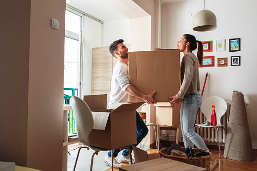 A couple moves a large cardboard box in a bright, modern living room, surrounded by packed items. They appear focused and determined.